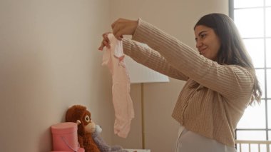 Huzurlu yatak odası ortamında bebek kıyafetlerine hayran bir kadın, sıcacık dcor ve doğal ışıkla sıcaklığını ve hazırlığını gösteren, beşikte gülümseyen İspanyol esmer.. 