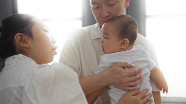 Baba bebeğini kucağında tutarken, anne sevgi dolu bir şekilde oturma odası düzenine bakıyor, aile bağını ve ev ortamını ön plana çıkarıyor..