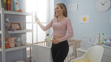 Yatak odasında duran bir kadın dekorasyon ve bebek beşiği ile gülümseyen bir selfie çekiyor, modern iç ve pozitif atmosfer sergiliyor..