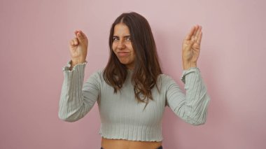 Pembe duvarın önünde elleri havada komik bir ifade sergileyen kadın, neşeli ve kendinden emin görünüyor, nazik ışıklandırmayla izole edilmiş, esmer saçlarını ve günlük kıyafetlerini vurguluyor..