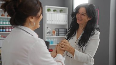 Klinik odasında gülümseyen, içten bir konuşma yapan kadınlar, arka planda tıbbi malzeme raflarıyla güven ve bakım göstergesi olarak, sıcak bir doktor-hasta etkileşimi öneriyorlar..