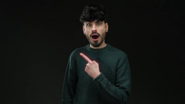 Yeşil kazaklı genç adam, şaşkın bir ifadeyle karanlık arka plana işaret ediyor merak ve entrika taşıyor..