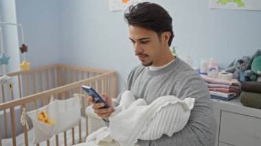 Genç adam kucağında bebek varken akıllı telefon kullanıyor, rahat ve huzurlu bir ev ortamı sergiliyor, aile bağlarını ve modern teknoloji kullanımını vurguluyor..