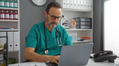 Tıbbi önlük ve gözlük takmış, dizüstü bilgisayara odaklanmış, boynunda steteskopla hastane kliniğinde çalışan İspanyol bir adam..