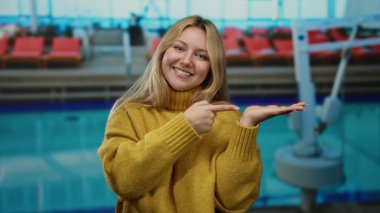 Havuz başında gülümseyen bir kadın açık eli işaret ediyor sarı süveter giyiyor açık havada rahat bir atmosfer..