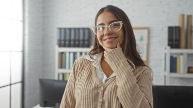 Arka planında kitap rafları olan bir ofis ortamında güvenle gülümseyen genç bir kadın, profesyonellik ve samimiyetle modern bir iş yeri havası sergiliyor..