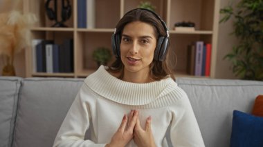 Kulaklık takan bir kadın, sıcak ve davetkar bir atmosferle çevrili göğsünde dinlenirken, güvenli bir şekilde gülümseyen modern dekorla rahat bir oturma odasında oturuyor..