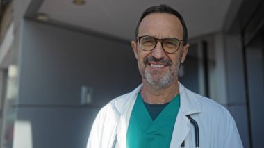 Şehir ortamında gülümseyen, gözlüklü ve tıbbi üniformalı olgun İspanyol bir adam profesyonel güven ve arkadaşlığı öneriyor..