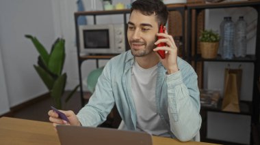 Ofisteki genç adam elinde bilgisayarlı, raflı ve arka planda bulunan kredi kartıyla telefonla konuşuyor..