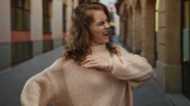 Kazaklı bir kadın şehir caddesinde acıdan suratını buruşturuyor. Rahatsızlığını ifade ediyor ve gergin bir ifadeyle omzunu tutuyor. Şehir hayatını ve sağlık sorunlarını gösteriyor..