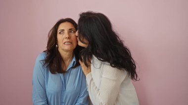 Pembe duvarda kız kardeşler fısıldıyor; iki orta yaşlı kadın mahrem anı paylaşıyor, sevgi, dostluk, sürpriz ve bağlantı paylaşıyorlar izole arka planda.