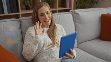 Kadın, sıcak bir oturma odasında rahat gri bir kanepede oturmuş kameraya doğru el sallarken ve sırıtırken evde tablet kullanarak video konferans yapıyor.