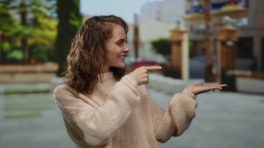 Genç bir kadın, açık hava parkında uzanan avuç içini göstererek gülümsüyor, rahat bir kazak giyiyor, güneşli günün tadını çıkarıyor ve bulanık yeşillik ve kentsel unsurlarla çevrili..