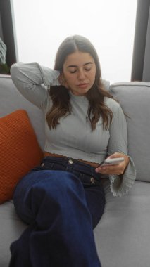 Modern bir oturma odasında, bir kanepede dinlenen, akıllı bir telefon tutan, bir ev ortamında gelişigüzel giyinen, dingin bir ifadeyle genç bir güzellik sergileyen bir kadın..