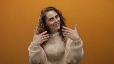 Turuncu arka planda gülümseyen bir kadın, rahat bir süveter içinde davetkar ve sıcak bir ortam sunan jestler yapar..