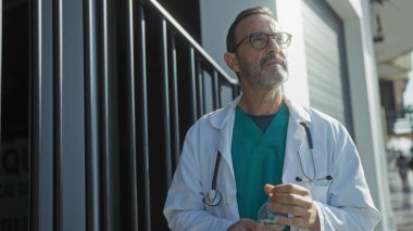 İspanyol doktor şehir ortamında beyaz önlük ve steteskop giyerek dikiliyor. Arka planda şehir binaları görünürken düşünceli görünüyor..