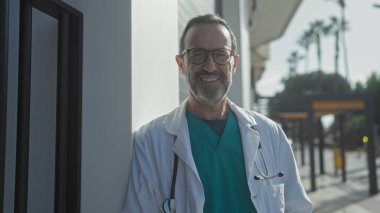 Kıdemli İspanyol doktor şehir ortamında gözlük ve önlükler takıp güneşli gökyüzünün altında steteskopla kameraya gülümsüyor..