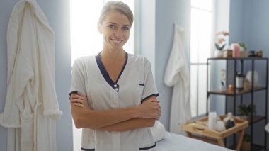 Klinikte kollarını kavuşturarak gülümseyen, üniforma giyen, özgüven ve sıcaklık saçan, hastanenin iç kısımlarıyla çevrili, profesyonellik ve özen gösteren bir kadın..