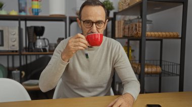 Orta yaşlı gözlüklü bir adam modern ofis içinde kahve içiyor. Mutfak elementleriyle tasarlanmış, konsantrasyon ve rahatlamayı yansıtan şık bir çalışma alanı sergiliyor..