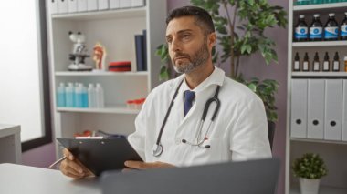 Tıbbi teçhizat ve raflarla çevrili, bir klinik odasında elinde bir pano tutan İspanyol erkek doktor, düşünceli bir şekilde beyaz önlük giyen tıbbi kayıtları inceliyor..