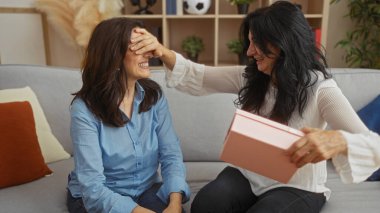 Kadınlar sevinçli bir anı paylaşırken biri diğerini şaşırtıyor. Sıcak bir oturma odası düzeniyle birlikte bağlarını ve mutluluklarını kutluyorlar..