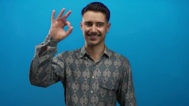 Şablonlu genç İspanyol adam canlı mavi arka planda, kendine güveni tam bir jest yaparak pozitiflik ve güvenilirliği ima ediyor..