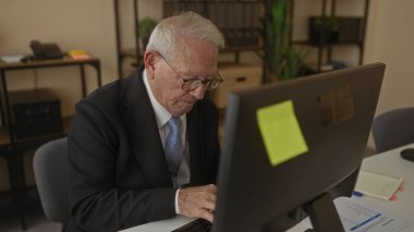 Ofiste bilgisayar başında çalışan kıdemli bir adam, akıllı telefonunu kontrol ediyor, etrafı kitaplar, bitkiler ve kırtasiye malzemeleriyle çevrili, profesyonel bir iş yeri düzeneği kuruyor..