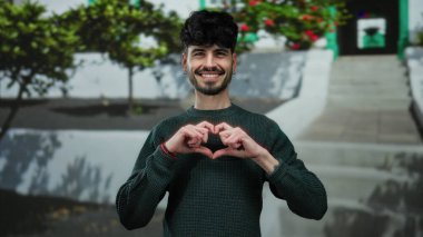 Bir kalp sembolü yapan genç bir adam şehir dışında renkli bir sanatsal arka plana karşı gülümseyen eller ile.