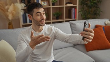 Genç adam, renkli yastıklar ve modern dekorlarla çevrili, rahat ve çekici bir atmosfer yayarak, rahat bir oturma odasında akıllı telefondan sohbet ediyor..