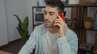 Genç İspanyol bir adam ofis ortamında modern dekorla telefonda konuşuyor, günlük kıyafetler giyiyor, odaklanmış ve özenli, profesyonel bir tavır sergiliyor..