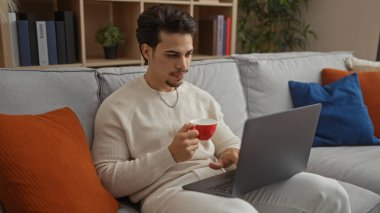 Genç adam rahat bir oturma odasında kanepede oturuyor, elinde kırmızı bardakla dizüstü bilgisayarla çalışıyor, rahat ve modern bir ev ortamını gösteriyor..