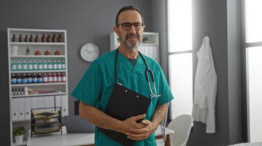 Steteskoplu ve dosyasıyla önlüklü İspanyol erkek doktor tıbbi malzeme ve ekipmanlarla dolu bir hastane odasında güvenle duruyor..