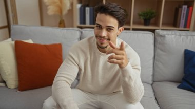Yakışıklı genç bir adam, rahat bir oturma odasında otururken kamerayı işaret ederek, kendine güvenen ve yaklaşılabilir bir tavır sergiliyor, sıcak dekorla iç mekanı vurguluyor..