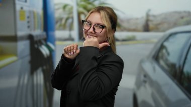 Sarışın ve gülümseyen bir kadın, başparmakları park halindeki bir arabanın yanındaki benzin istasyonuna dönük bir şekilde güvenli bir şekilde poz veriyor, açık hava kentsel hayatının bir anını yakalıyor..