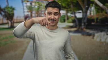 Parkta gülümseyen genç İspanyol bir adam. Arka planda ağaçlar ve patika var. Açık mavi gökyüzünün altında doğal çevreye güveni var..