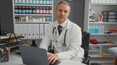 Hastane odasında dizüstü bilgisayarla çalışan sarı saçlı ve steteskoplu olgun bir adam klinik iç mekanda çalışan bir tıp uzmanını öneriyor..