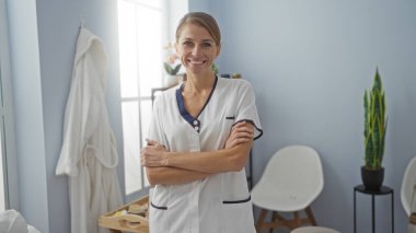 İçerideki bir klinikte beyaz bir üniforma içinde gülümseyen ve kolları pencerenin ve bitkilerin yanında çapraz duran bir kadın sakin bir ortamda kendine güven ve profesyonellik yayıyor..