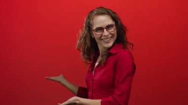 Kırmızı bluzlu ve gözlüklü gülümseyen genç bir kadın canlı kırmızı bir arka plana uzanmış ellerini uzatarak izole bir ortamda neşe ve coşku taşıyor..