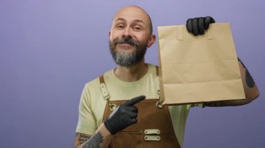 Sakallı, eldiven ve önlük takan kel bir adam, elinde kese kağıdıyla, izole edilmiş mor arka planda rahatça poz veriyor..