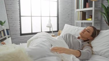 Güneşli yatak odasında uyanan kadın rahat bir yatakta rahat bir dekor ve yumuşak yastıkla mutlu bir şekilde uzanıyor..