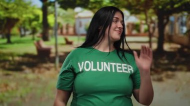 Yeşil tişörtlü genç bir kadın parkın dışında gülümsüyor ve sosyal farkındalık aktivitelerine katılıyor..