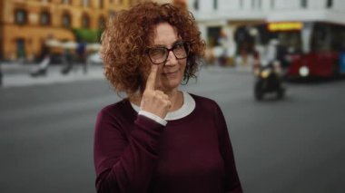 Şehir caddesinde duran kadın, arka planda bulanık otobüsle gözü işaret ederek merak ve şehir yaşamını işaret ediyor..