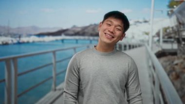 Deniz kenarındaki gezinti alanında gülümseyen ve gözlerini ovuşturan, günlük giysiler giyen, manzaralı bir plaj ve arka planda açık mavi gökyüzü olan genç bir adam..