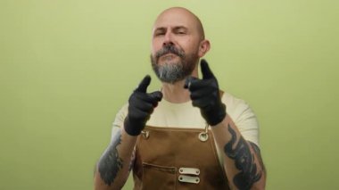 Kel, sakallı, eldivenli, yeşil bir duvara bakan, kahverengi önlüklü, elinde dövmeleri olan bir adam.