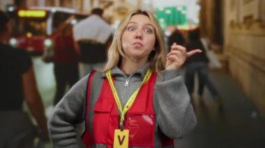 Kırmızı yelek giyen kadın şehir caddesinde, bulanık yayalar ve araçlarla çevrili, gönüllü şehir çalışmalarını gösteren hareketli bir jest yapıyor..