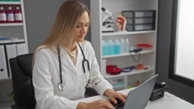 Klinikte steteskop takan bir kadın ofiste dizüstü bilgisayarla çalışırken, gün içinde hastanede tıbbi ekipman ve belgelerle çevrili bir şekilde çalışıyor..