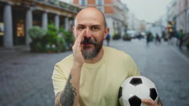 Kentsel sokak ortamında futbol topu tutan, sakallı, kel, dövmeli ve şehir ortamında odaklanmış arkadaşça ifade veren gizli fısıldayan bir adam..