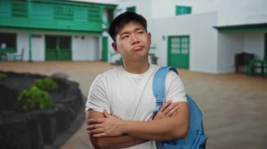 Sırt çantalı genç Çinli adam şehir caddesinde çapraz kollu duruyor, açık havada binaların ve yeşilliklerin yanında beyaz gömlek giyiyor, düşünceli görünüyor.