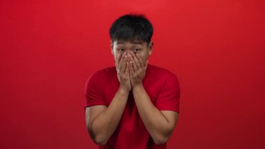 Şaşırmış ifadesi olan genç bir adam, izole bir arka planda parlak kırmızı bir duvara karşı elleri ile ağzını kapatır.
