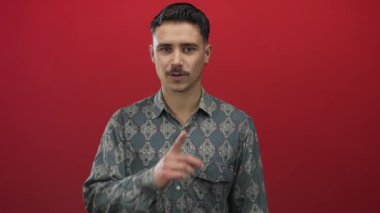 Genç ispanyol adam sessiz hareketlerle dudakları üzerinde canlı kırmızı arka planda gizlilik ve sakinliği öneriyor..